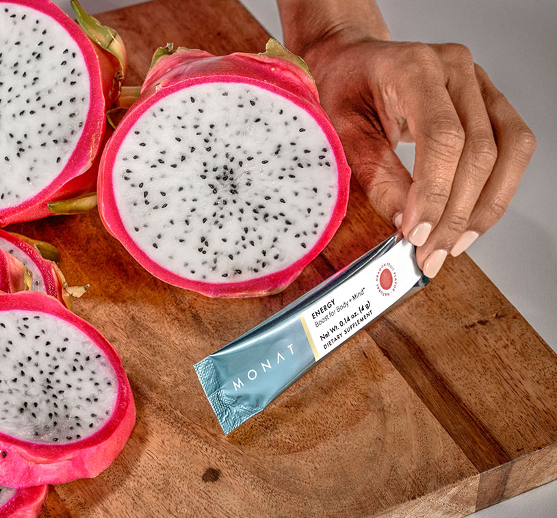 Fresh cut dragon fruit sitting on a wooden cutting board, next to a hand holding a MONAT Energy™ Natural Dragon Fruit stick pack.