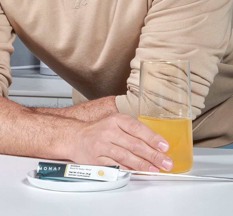 Male hand holding a glass of water mixed in with MONAT Energy™ Natural Pineapple, next to a MONAT Energy™ Natural Pineapple stick pack.