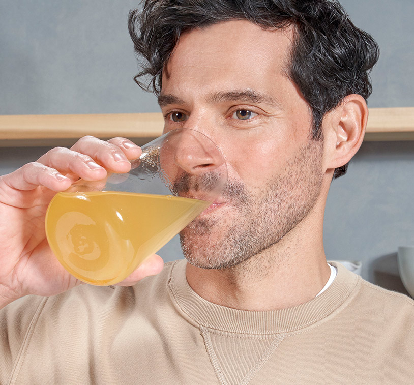 Brunette male drinking MONAT Energy™ Natural Pineapple out of a glass cup, overlayed on an image of fresh pineapple.