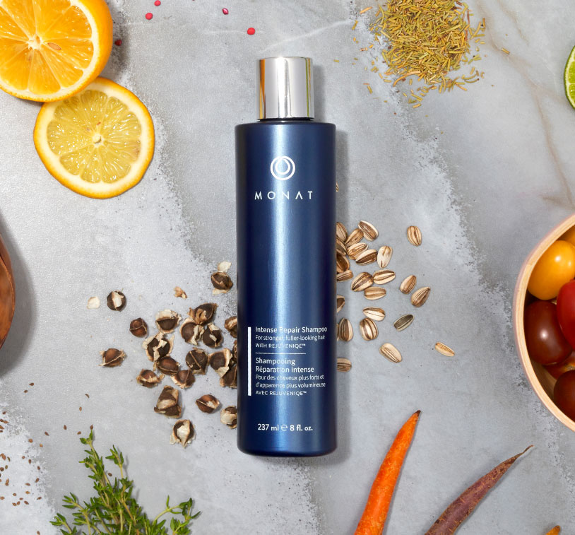 MONAT’s Damage Repair Shampoo stands over a white solid background setting displaying raw ingredients such as oils, citrus & 
					herbs along with additional glass ornaments around it.