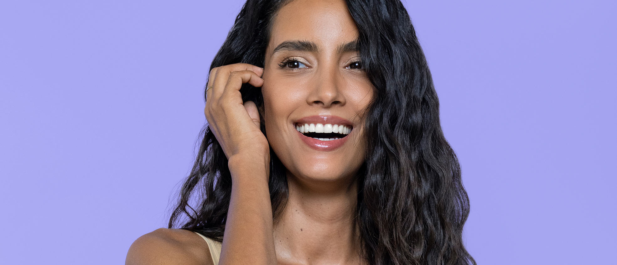 A smiling female with dark brown wavy hair, with her hand on her face, while standing infront of a purple background.