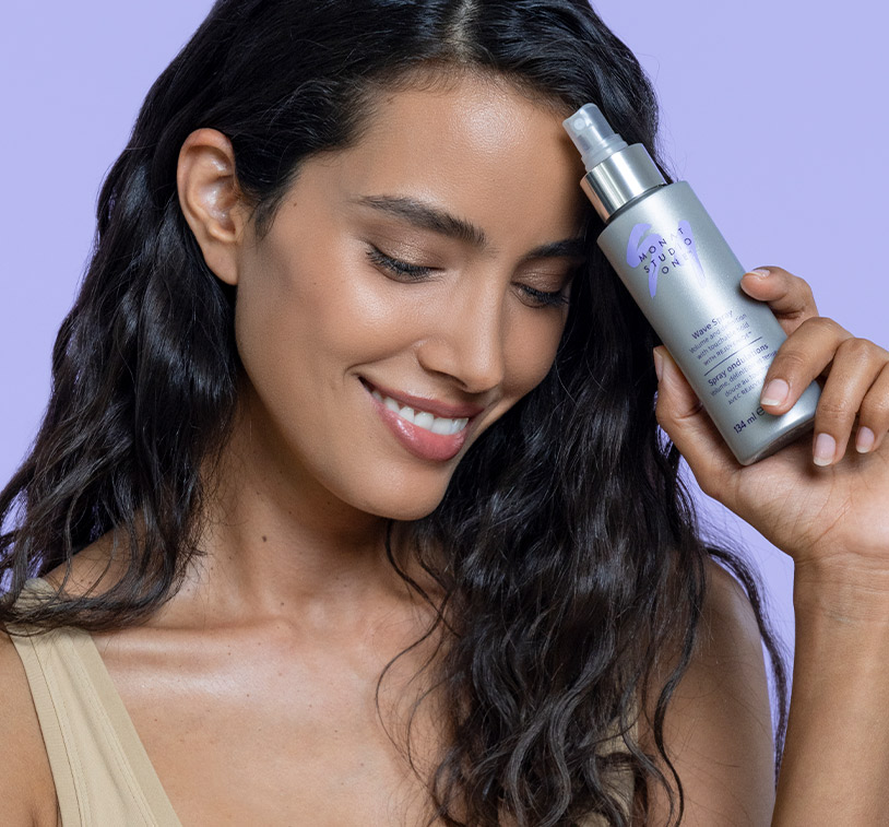 A female with dark brown wavy hair, smiling while holding MONAT STUDIO ONE™ Wave Spray, overlapping a photo of an open coconut and aloe vera.