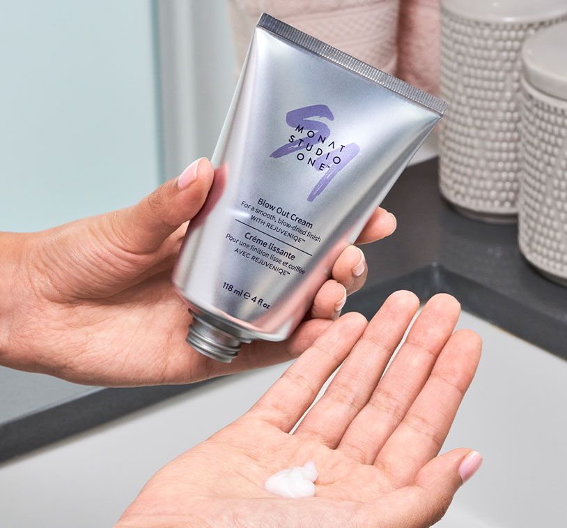 Female hand infront of a bathroom sink holding MONAT STUDIO ONE™ Blow Out Cream, while the other hand has product in it.