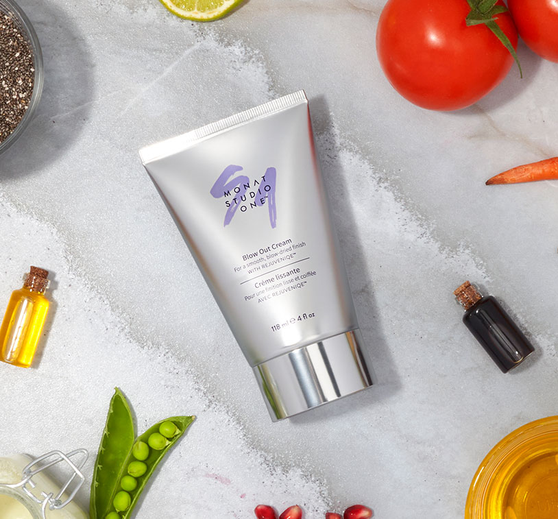 MONAT STUDIO ONE™ Blow Out Cream laying on marble tile surrounded by ingredients such as tomatos, limes, carrots and pomegranates.