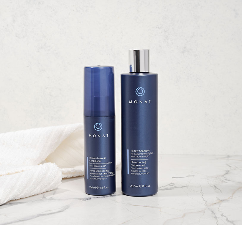 A hand grabbing MONAT STUDIO ONE™ The Moxie Magnifying Mousse on a white counter, next to gray bath towels and two glass containers.
