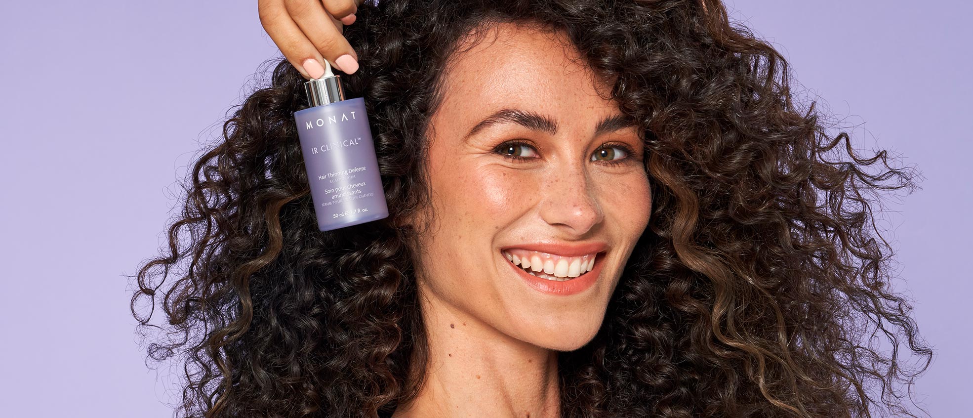 Female with brown curly hair smiling while holding IR Clinical™ Hair Thinning Defense.