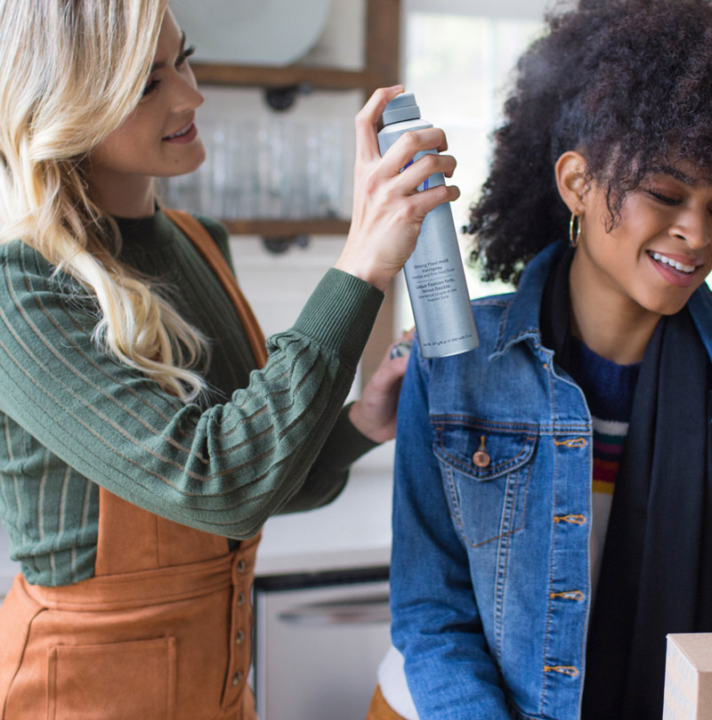 Blonde female spraying hairspray onto a brunette female's curly hair.