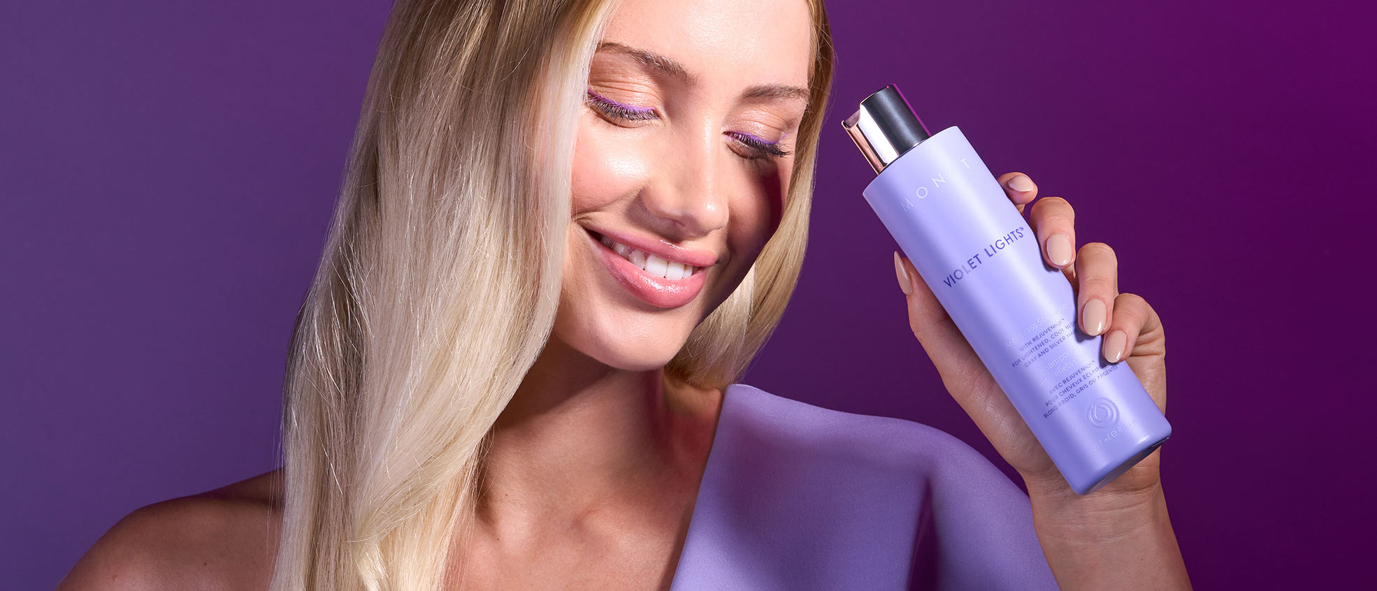 Blonde female smiling while holding Violet Lights™ Anti-Brass Shampoo in front of her face.