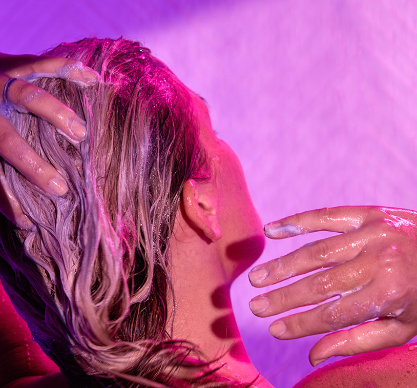 Blonde female washing her hair in the shower with Violet Lights™ Anti-Brass Toning Masque, while overlaying a texture shot of Violet Lights™ Anti-Brass Toning Masque.