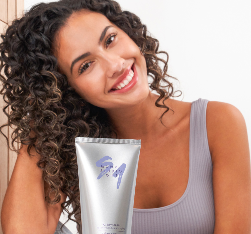 curly hair female poses while holding an air dry cream product in front of her.