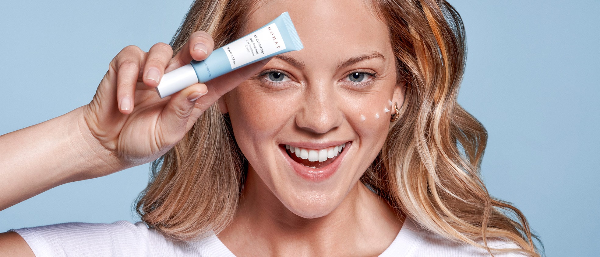 A blonde female smiling and holding BE CLARIFIED™ Spot Treatment infront of her face, while having a few drops of the treatment on her cheek.