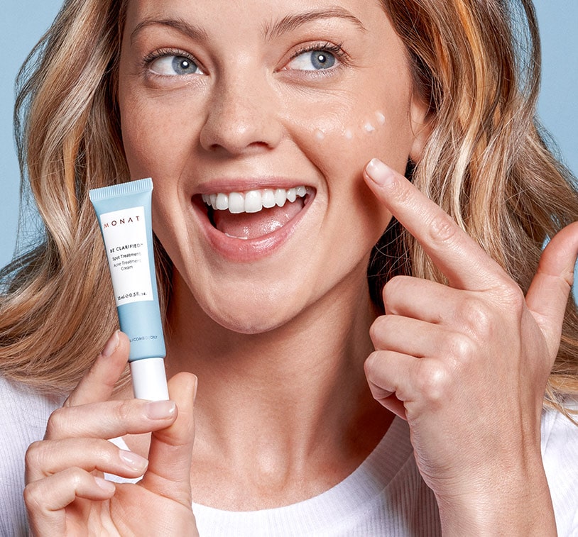 A blonde female smiling and holding BE CLARIFIED™ Spot Treatment infront of her face with a few drops of the treatment on her cheek, while overlaying a photo of 4 BE CLARIFIED™ Spot Treatments.