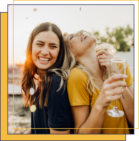 Brunette female and blonde female laughing while standing outside and drinking champagne.