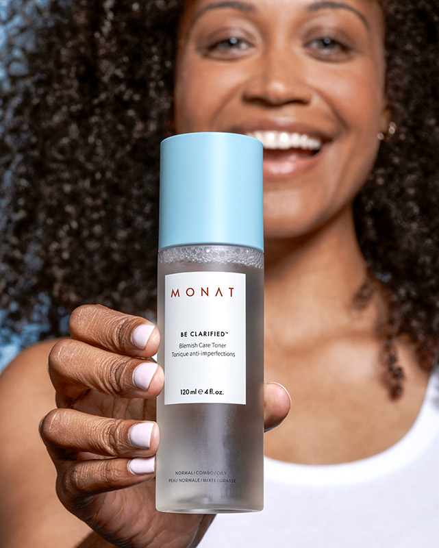 A brunette female with curly hair smiling while holding BE CLARIFIED™ Blemish Care Toner.