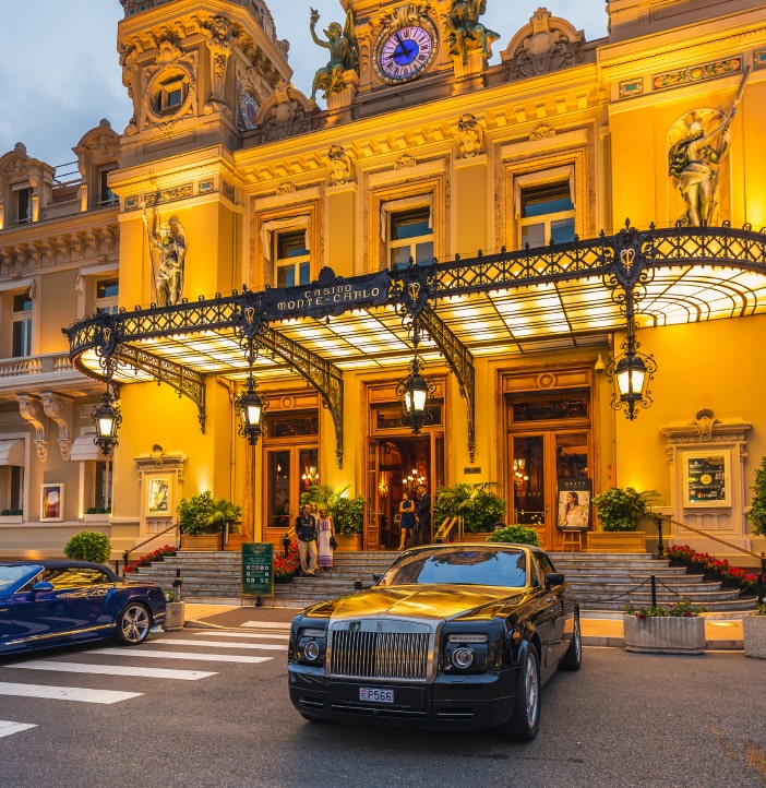 photo shot of a main facade of Monte Carlo Hotel with people in his entrance and a couple of cars parking here