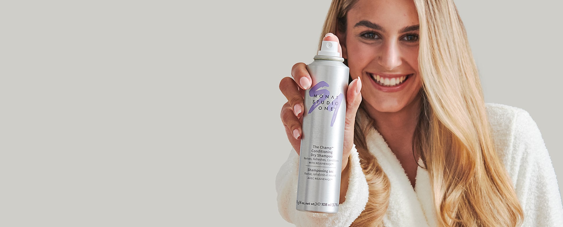  A blonde female smiling while holding the MONAT STUDIO ONE™ THE CHAMP™ Conditioning Dry Shampoo. 