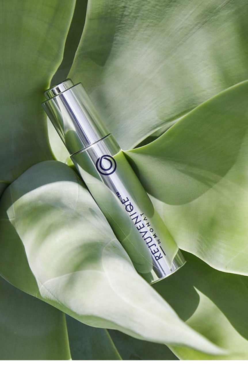 A silver Rejuveniqe oil bottle rests diagonally on overlapping green leaves, illuminated by soft natural light.