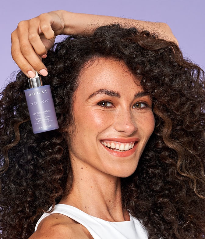 curly female holding an IR CLINICAL™ HAIR THINNING DEFENSE SCALP SERUM next to here face on a light pruple background