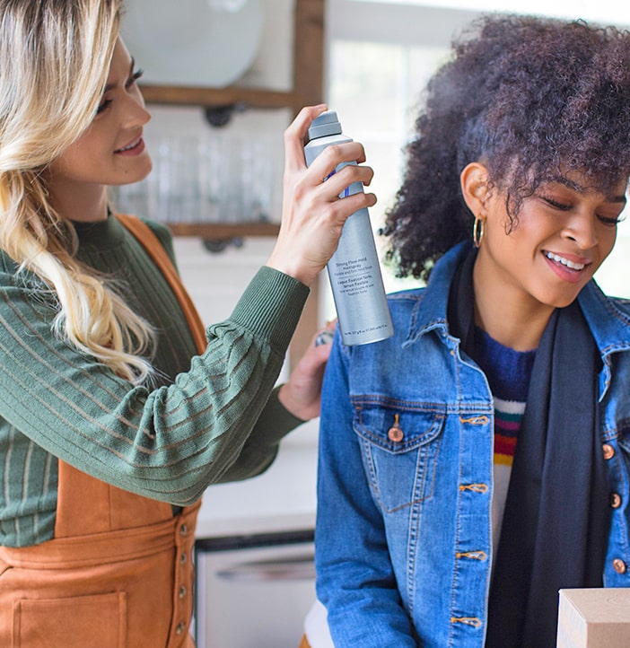 blonde female apliying a MONAT spray on a curly hair of a black woman