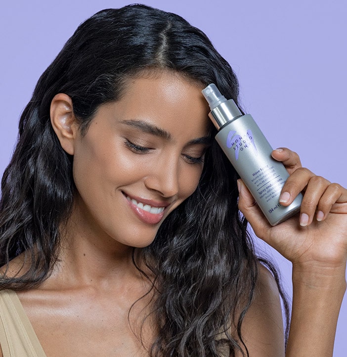 brunette female posing with a MONAT STUDIO ONE™ WAVE SPRAY next to her face on a light purple background