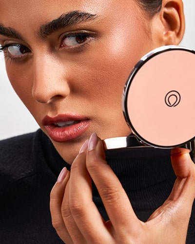 Female holding an MONAT Radiant Bronzer™, while looking in the mirror.