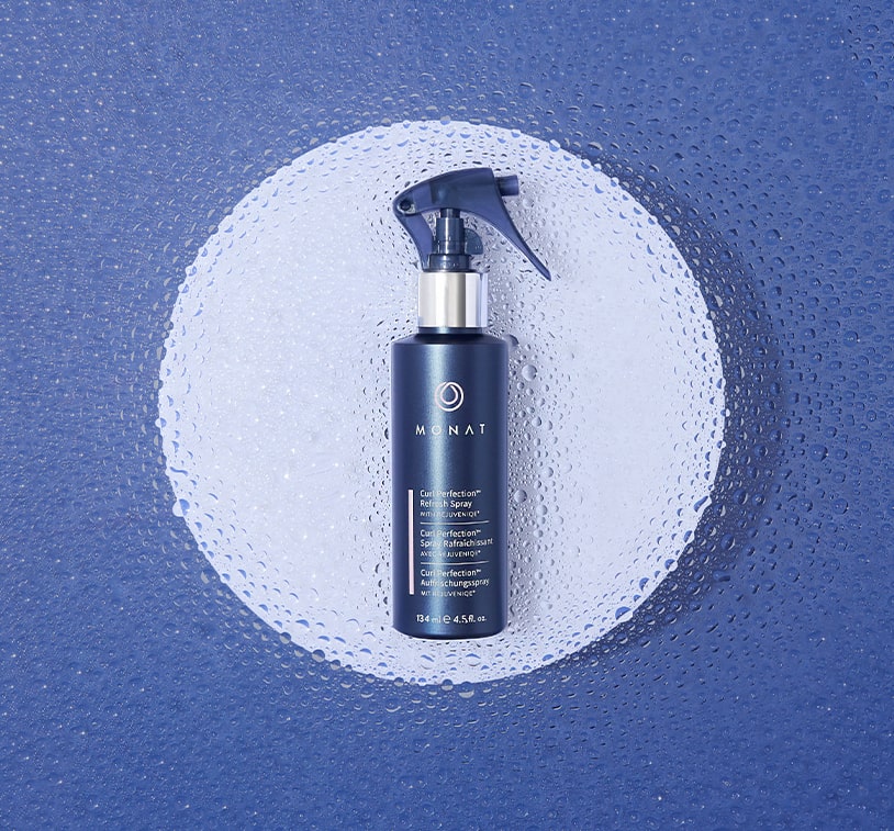 Image of a bottle of 'MONAT Curl Perfection™ Refresh Spray' lying on a blue and white surface with water droplets around it.