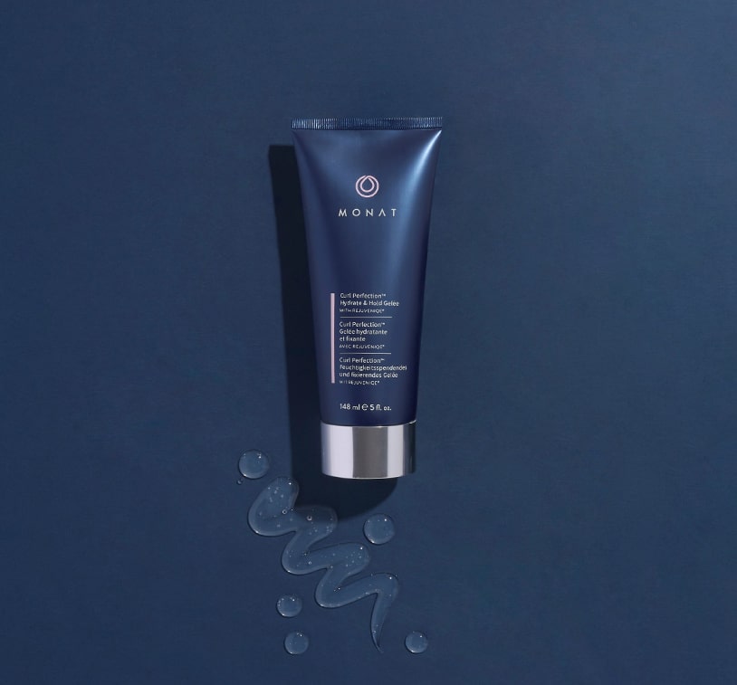 Image of a tube of 'MONAT Curl Perfecting Hydrate & Hold Gelée' lying on a blue surface with water droplets around it. 