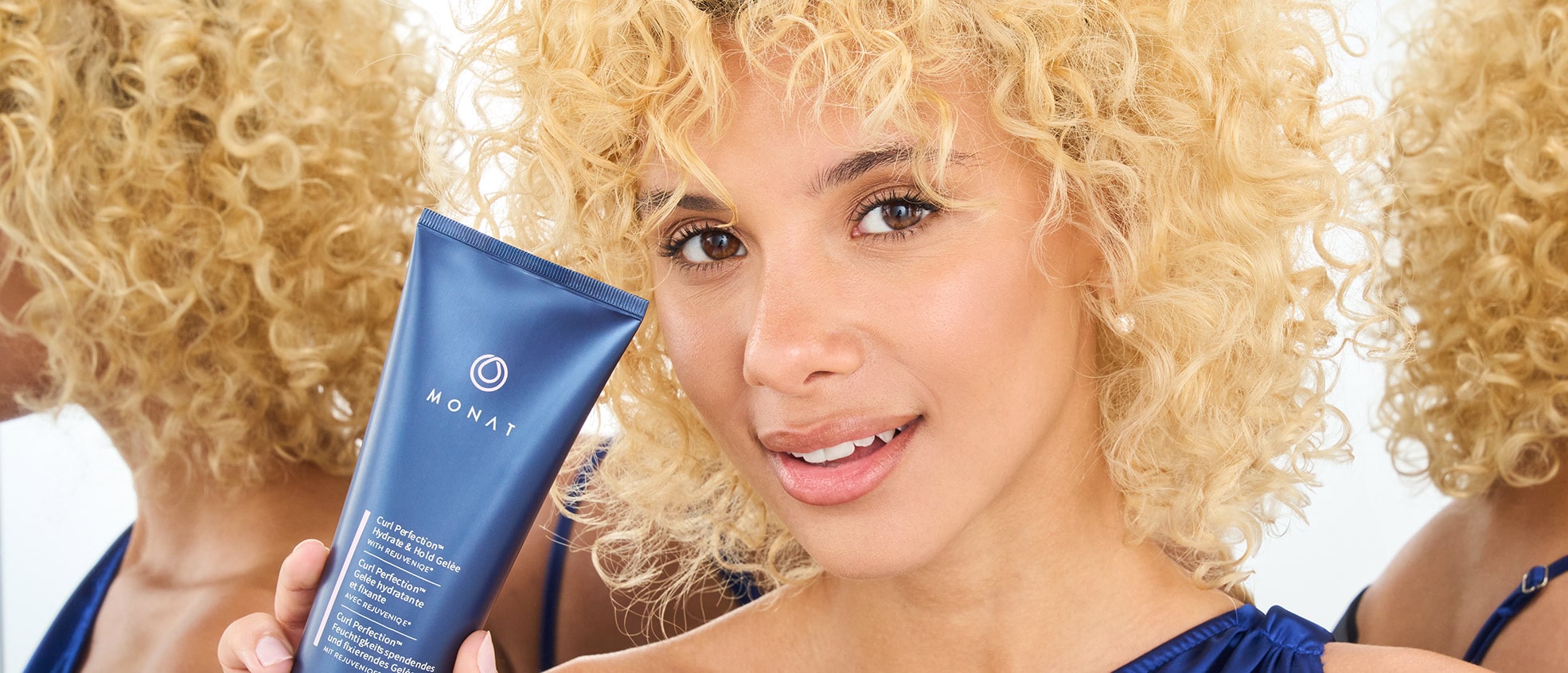 Image of a woman with curly blonde hair holding a tube of 'MONAT Curl Perfecting Hydrate & Hold Gelée' and smiling.