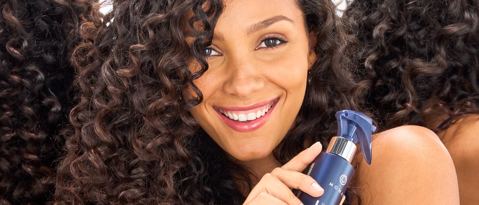 Smiling woman with curly hair holding a bottle of MONAT Curl Perfection™ Refresh Spray.