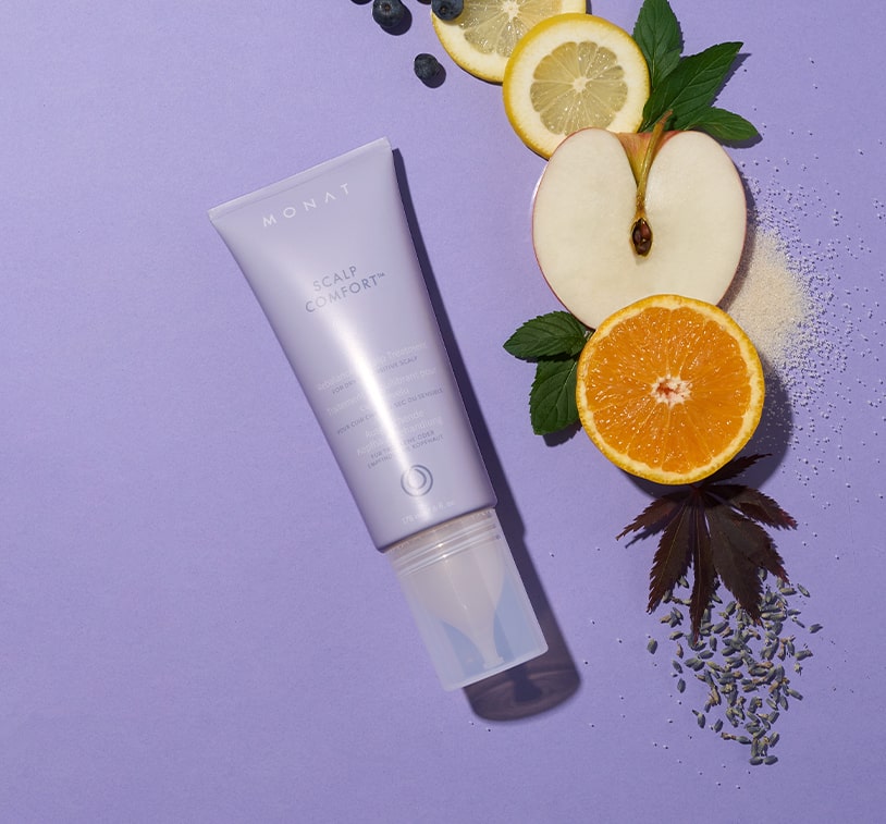 Scalp Comfort Rebalancing Serum bottle lying on a purple surface, surrounded by fresh fruit slices and botanical leaves.