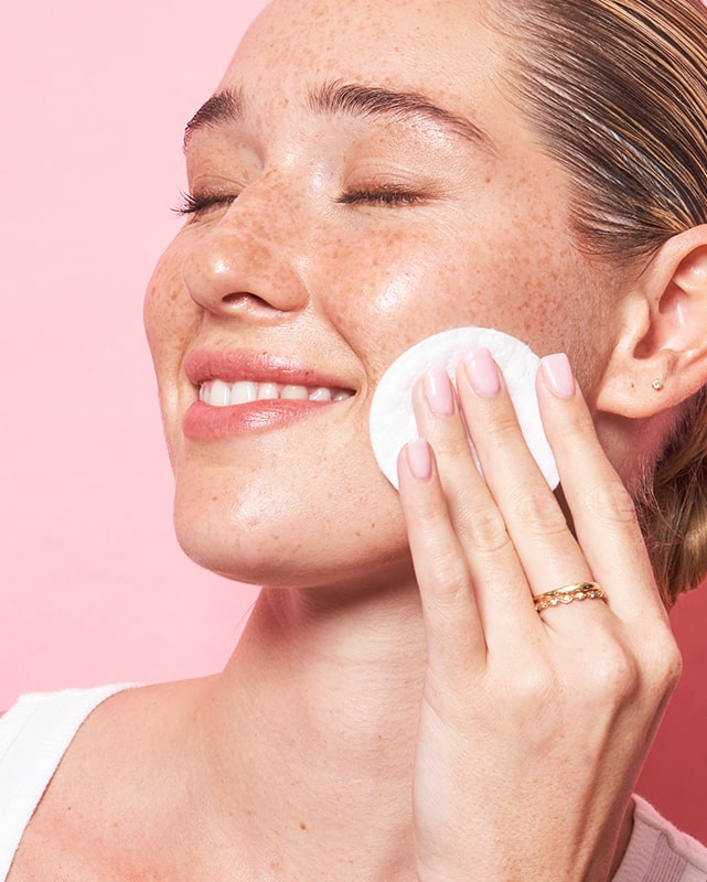 Smiling woman with clean skin using MONAT Micellar Water with a cotton pad on her face.