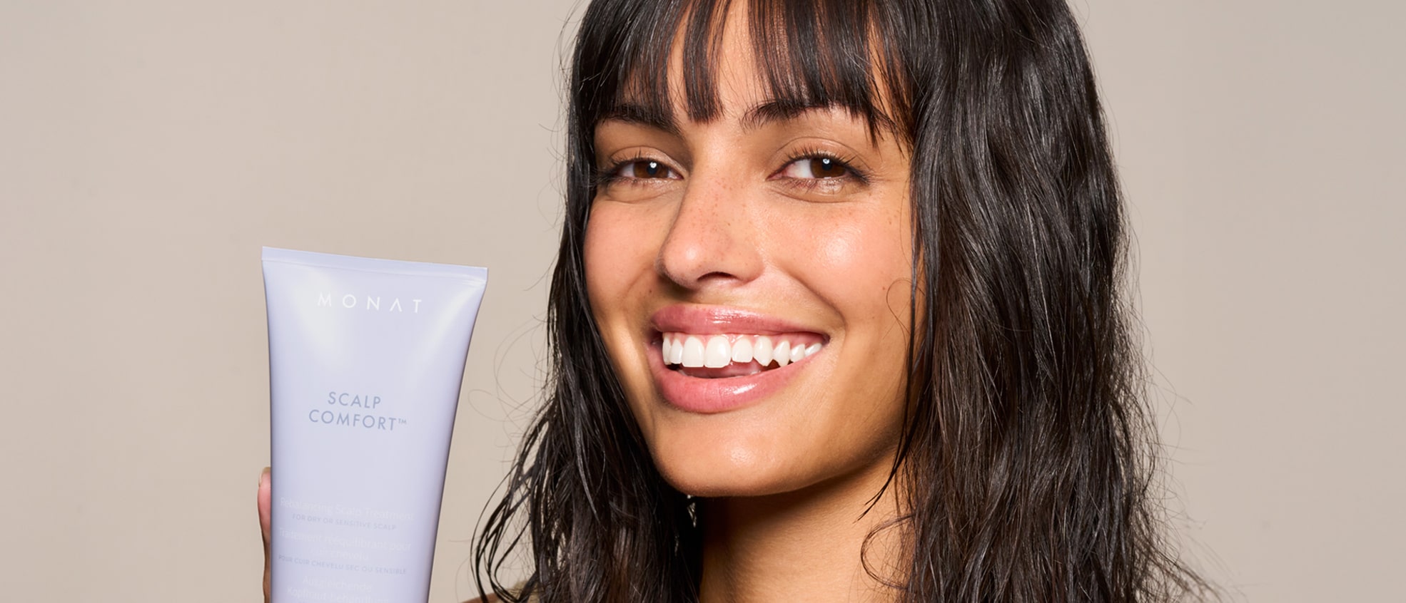 Close-up of a smiling model with dark hair holding the MONAT Scalp Comfort Treatment tube near her face.