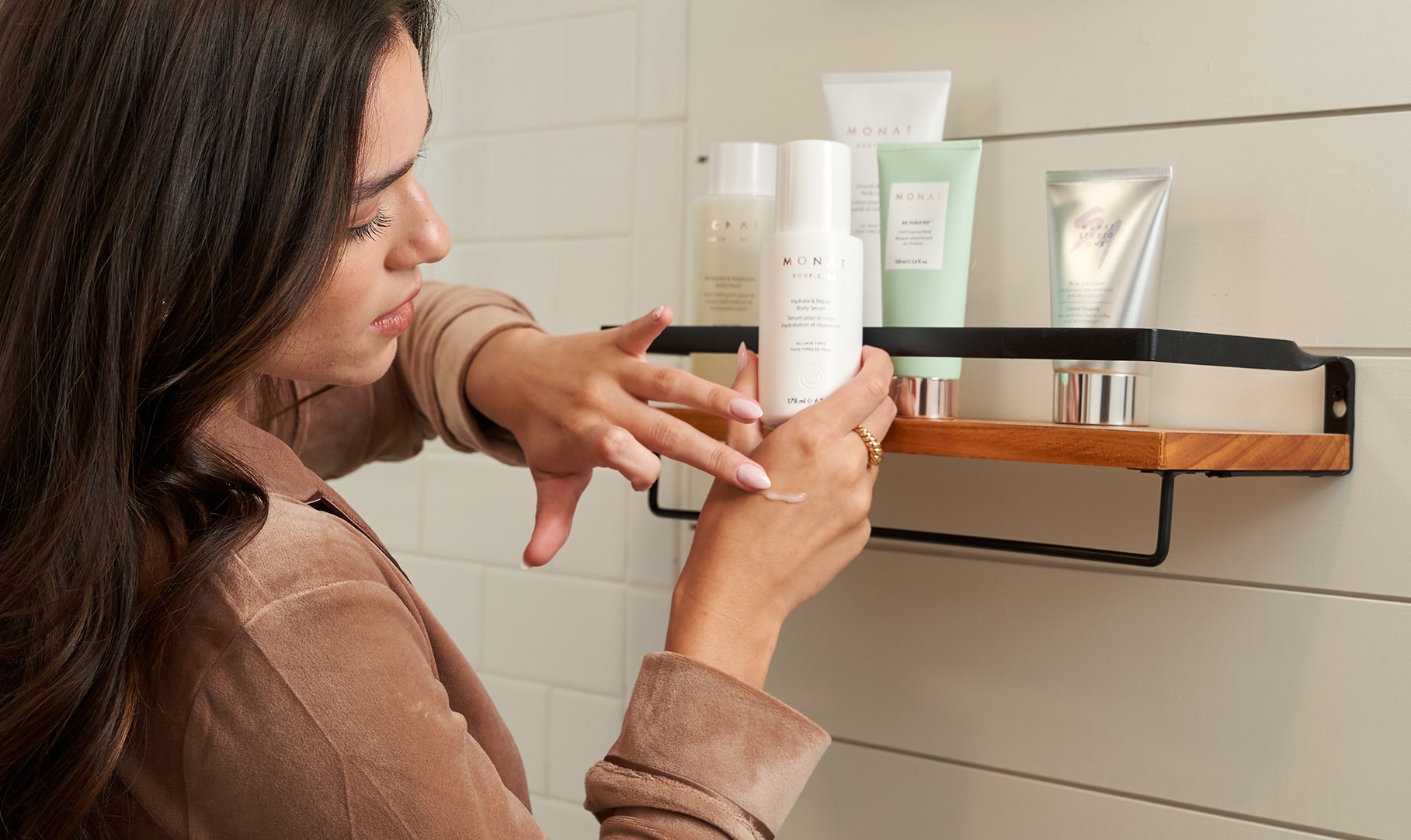 A dark-haired woman in a bathroom, smiling while reaching for a Monat product on a stylish shelf filled with other beauty products.