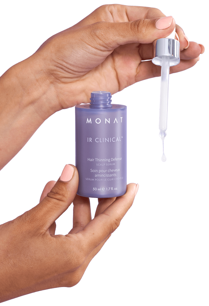 Hands holding a Monat IR Clinical Hair Thinning Defense serum bottle with a dropper releasing a drop of product.