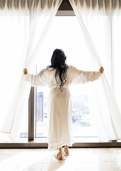 A woman in a bathrobe draws open the curtains of a luxurious suite