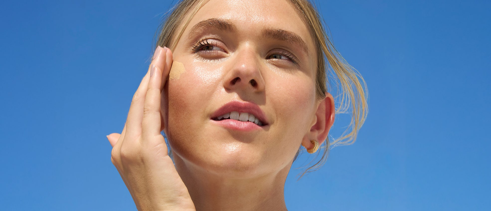 Close-up of a woman smiling and looking up under a clear blue sky, showcasing glowing skin.