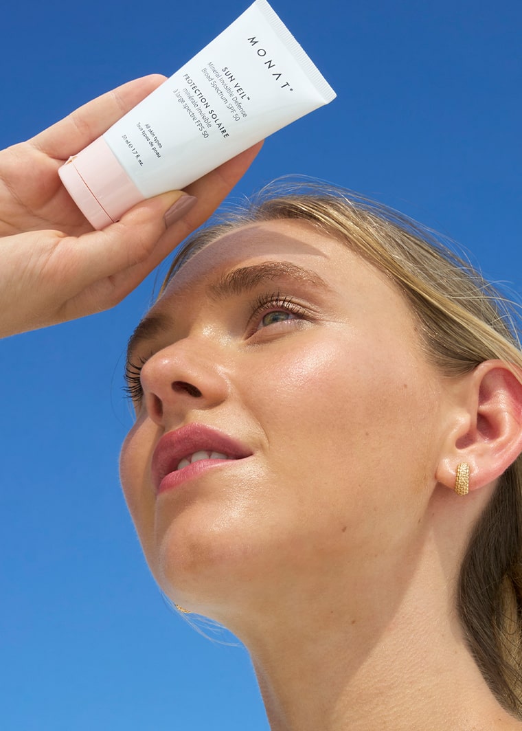 Woman wrapped in soft white fabric holding MONAT Sun Veil SPF 50 toward the camera, with blue sky in the background