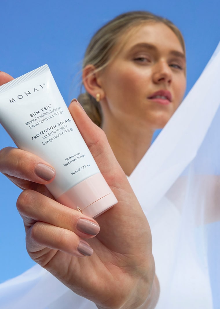 Woman wrapped in soft white fabric holding MONAT Sun Veil SPF 50 toward the camera, with blue sky in the background