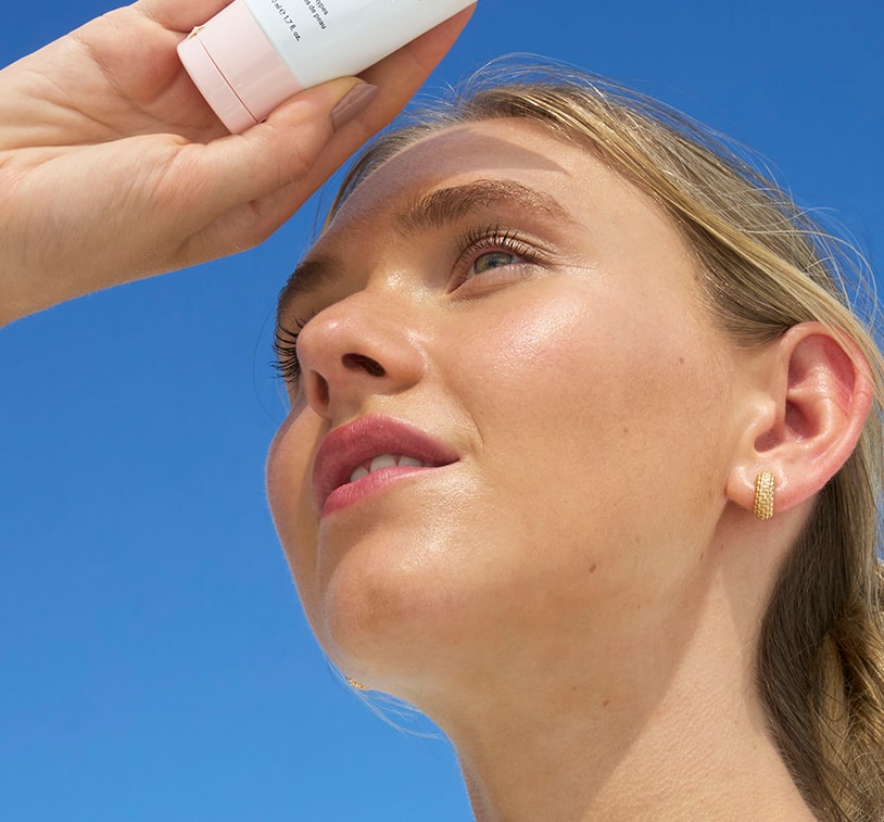 Smiling woman holding Sun Veil Mineral Invisible Defense-Broad Spectrum-SPF-50 tube near face, indoors with warm lighting.