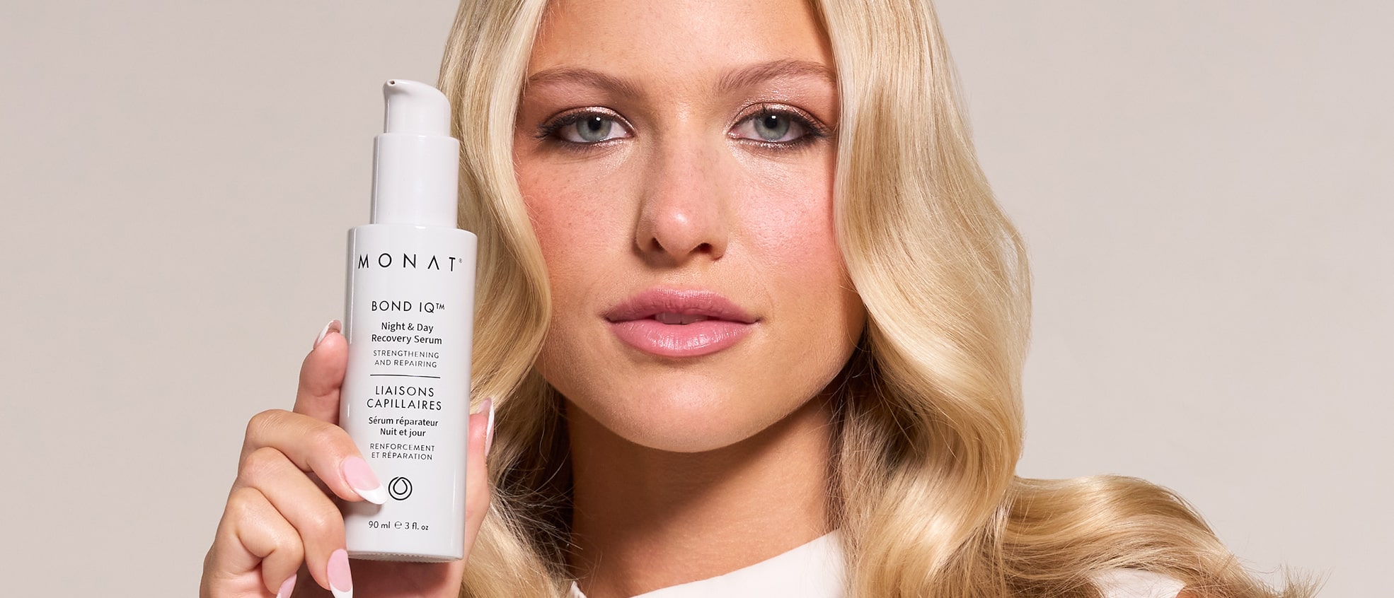 A blonde woman with wavy hair holds a white bottle of MONAT BOND IQ Night & Day Recovery Serum close to her face