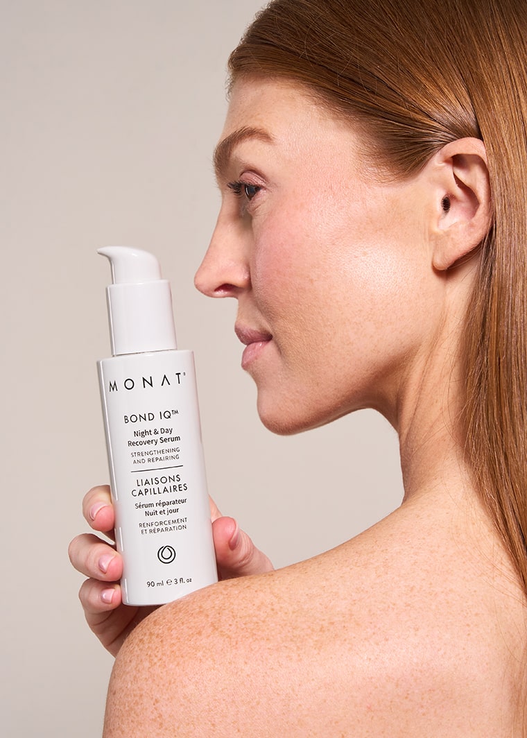 Profile view of a woman with straight, honey-blonde hair pulled back, holding a MONAT BOND IQ serum bottle