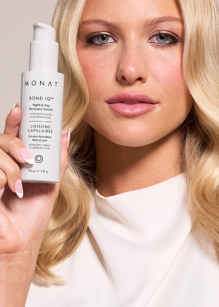 A blonde woman with wavy hair holds a white bottle of MONAT BOND IQ Night & Day Recovery Serum close to her face