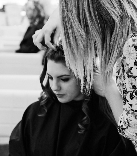 A black and white photograph showing a hair styling session, with one person working on another's hair