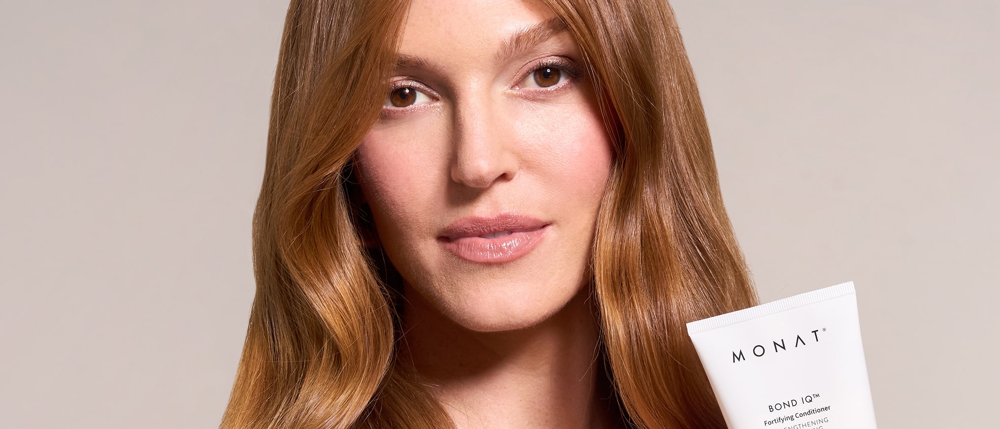 Close-up portrait of a woman with long, straight brown hair and brown eyes, holding a MONAT BOND IQ Fortifying Conditioner tube