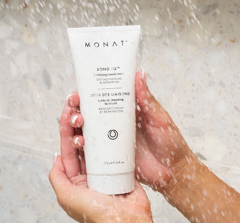 Hands holding a MONAT BOND IQ Fortifying Conditioner tube under running water in a shower