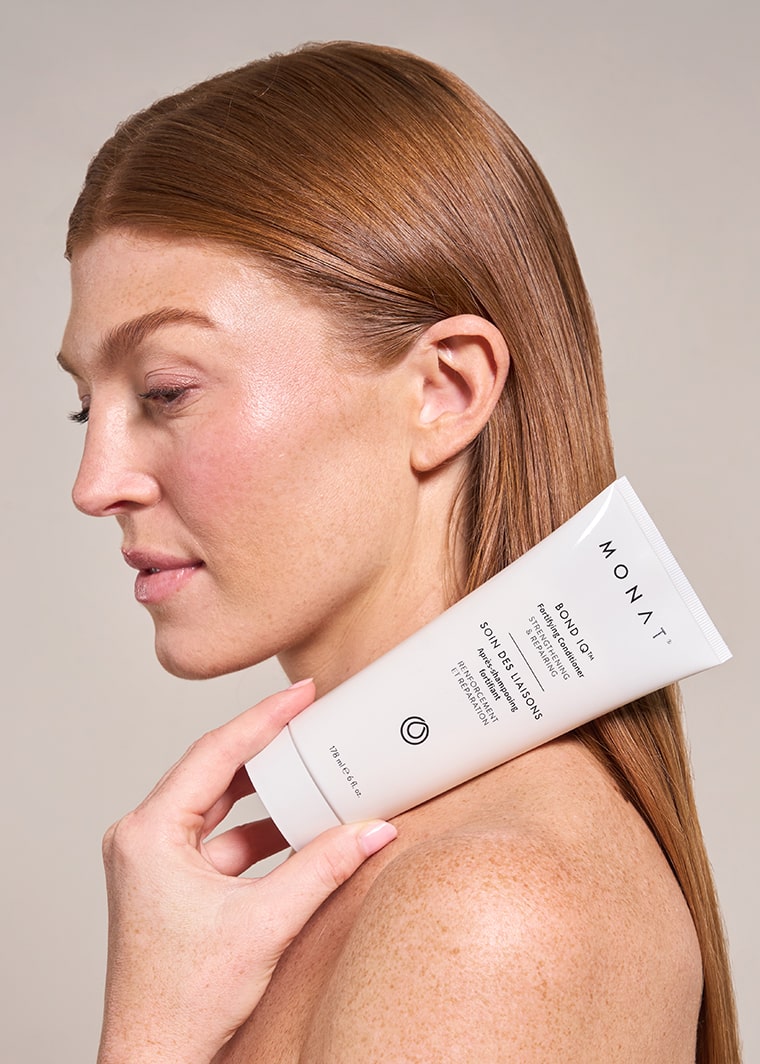 Profile view of a woman with sleek, straight auburn hair pulled back, holding a white tube of MONAT BOND IQ Fortifying Conditioner against her bare shoulder