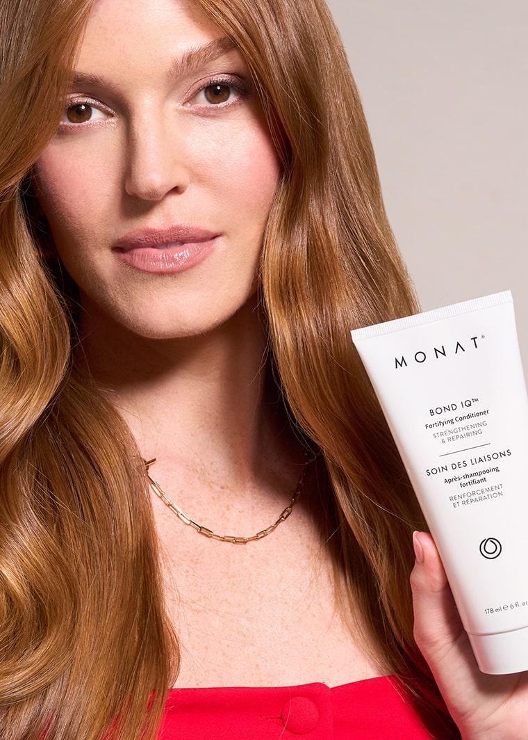 A woman with long, wavy brown hair and brown eyes holds a white MONAT BOND IQ Fortifying Conditioner tube