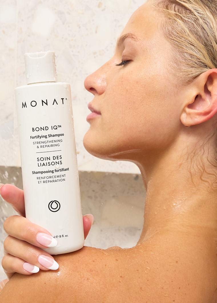Close-up of a woman in the shower holding a bottle of MONAT Bond IQ™ Fortifying Shampoo near her shoulder, with water droplets on her skin and the bottle