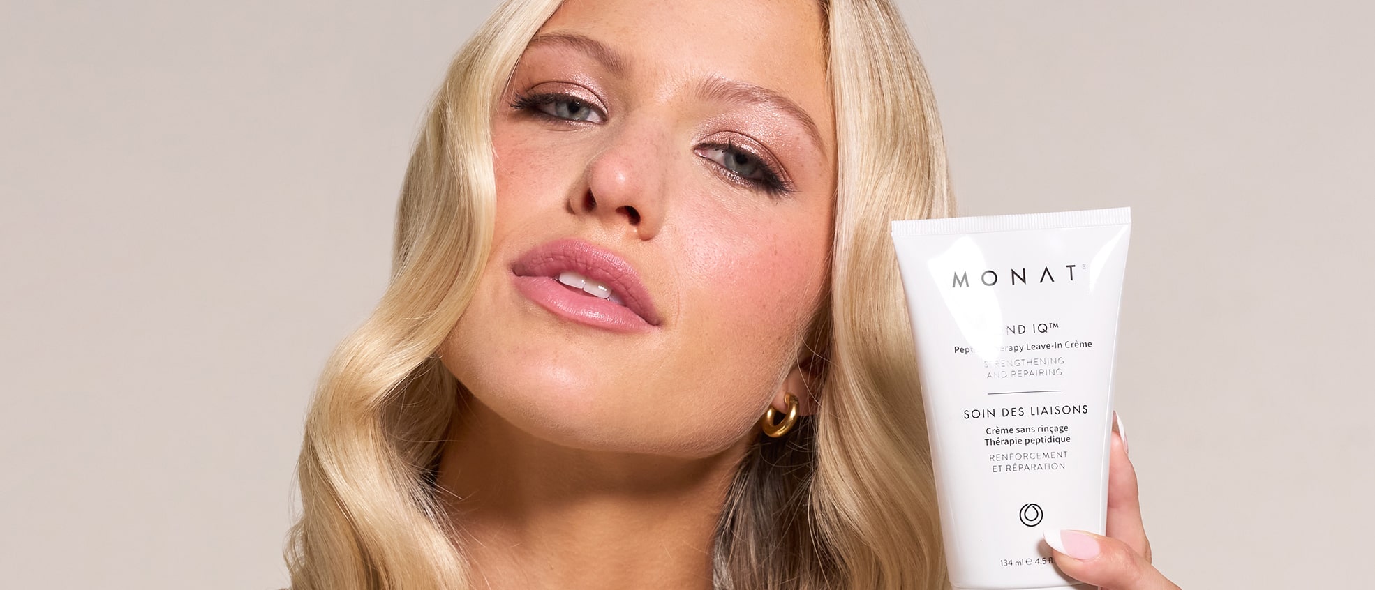 Close-up of a blonde woman with sleek hair holding the tube of crème near her face