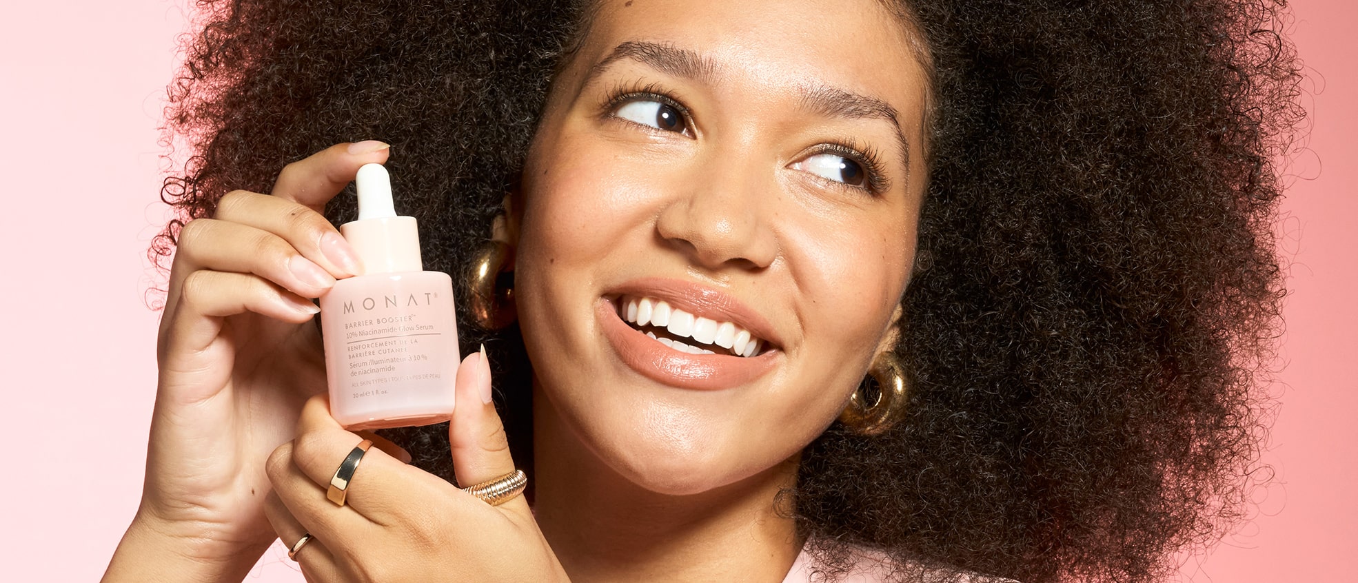 Smiling woman with curly hair holding MONAT Barrier Booster™ Niacinamide Glow Serum dropper bottle near her face.